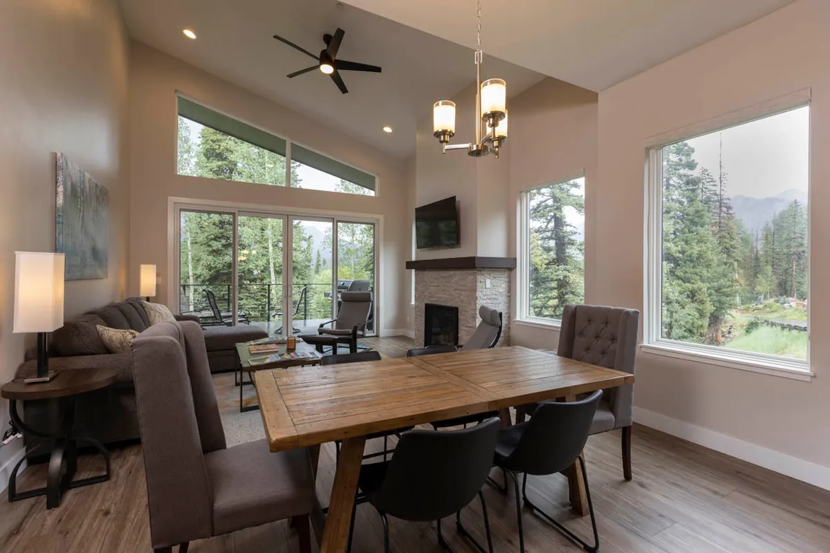 The image shows a cozy, open-concept living and dining area with a plush sofa, inviting dining table, and expansive windows framing breathtaking views of Colorado's mountains and forests. Sunlight pours in, illuminating the room and creating a warm, welcoming atmosphere—an ideal setting for family gatherings or quiet relaxation.