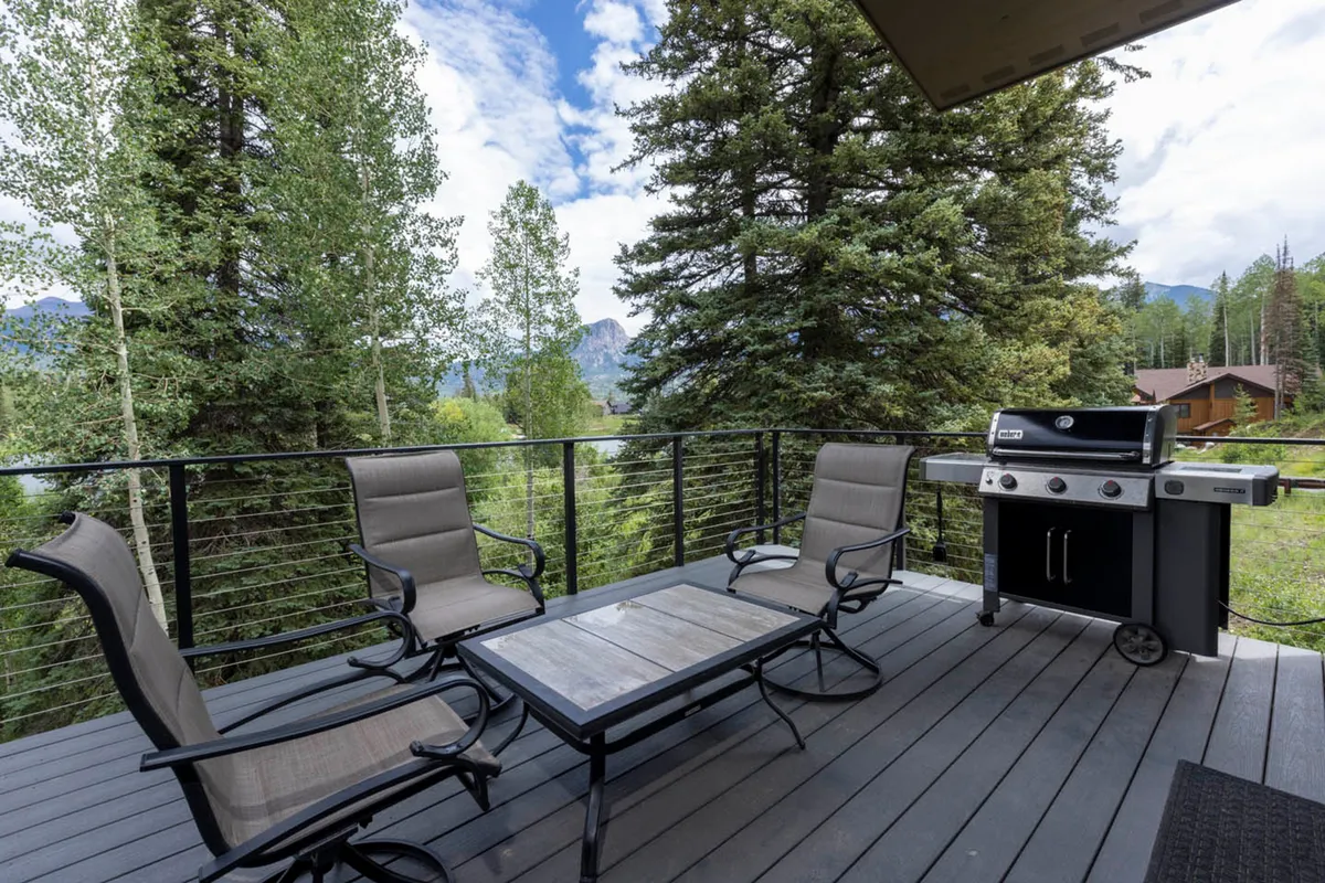 This image features a cozy outdoor seating area on a balcony or deck, complete with a table, grill, and comfortable chairs. The setup faces a stunning mountain view framed by surrounding trees, creating an inviting spot to relax and enjoy the scenery.