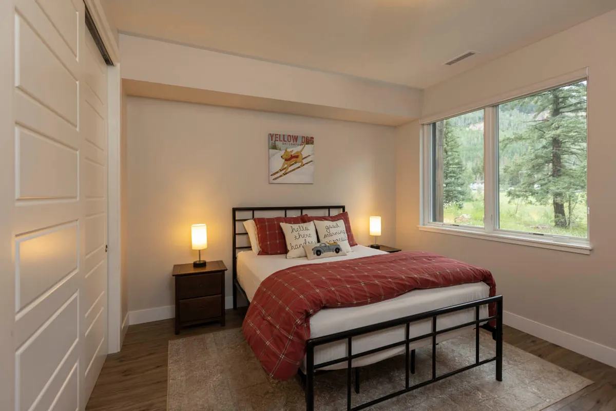 A comfortable bedroom featuring a king-size bed with a red plaid blanket, large window filling the room with natural light, nightstands with lamps, and playful dog-themed wall art. The inviting space exudes warmth and offers mountain views, emphasizing comfort and relaxation.