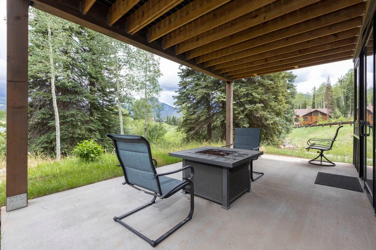 An inviting exterior scene featuring a fire pit surrounded by outdoor seating, with tall trees and distant mountain views providing a tranquil natural backdrop. The setting is perfect for families to gather, relax, or enjoy outdoor meals by the water’s edge.