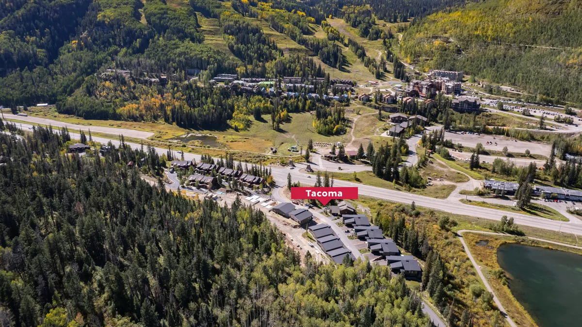 A scenic exterior shot featuring the Tacoma sign surrounded by lush greenery, with residential buildings and a roadway in the background. The area looks inviting for outdoor activities like hiking, biking, and leisurely walks, with a sense of community and access to nature.