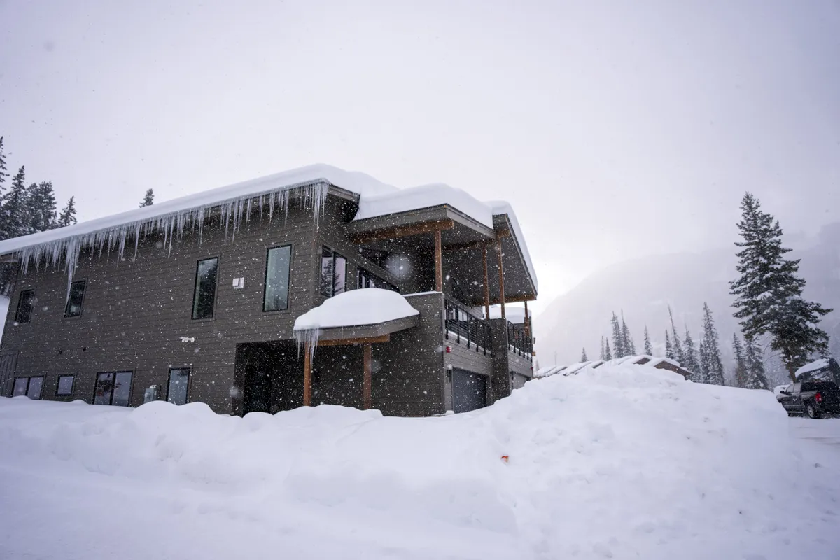 A modern home framed by snow-laden roofs, large windows, icicles, and stately pine trees. The setting suggests warm moments inside after days filled with winter activities like skiing and snowshoeing, while the exterior hints at the possibilities for outdoor excitement.