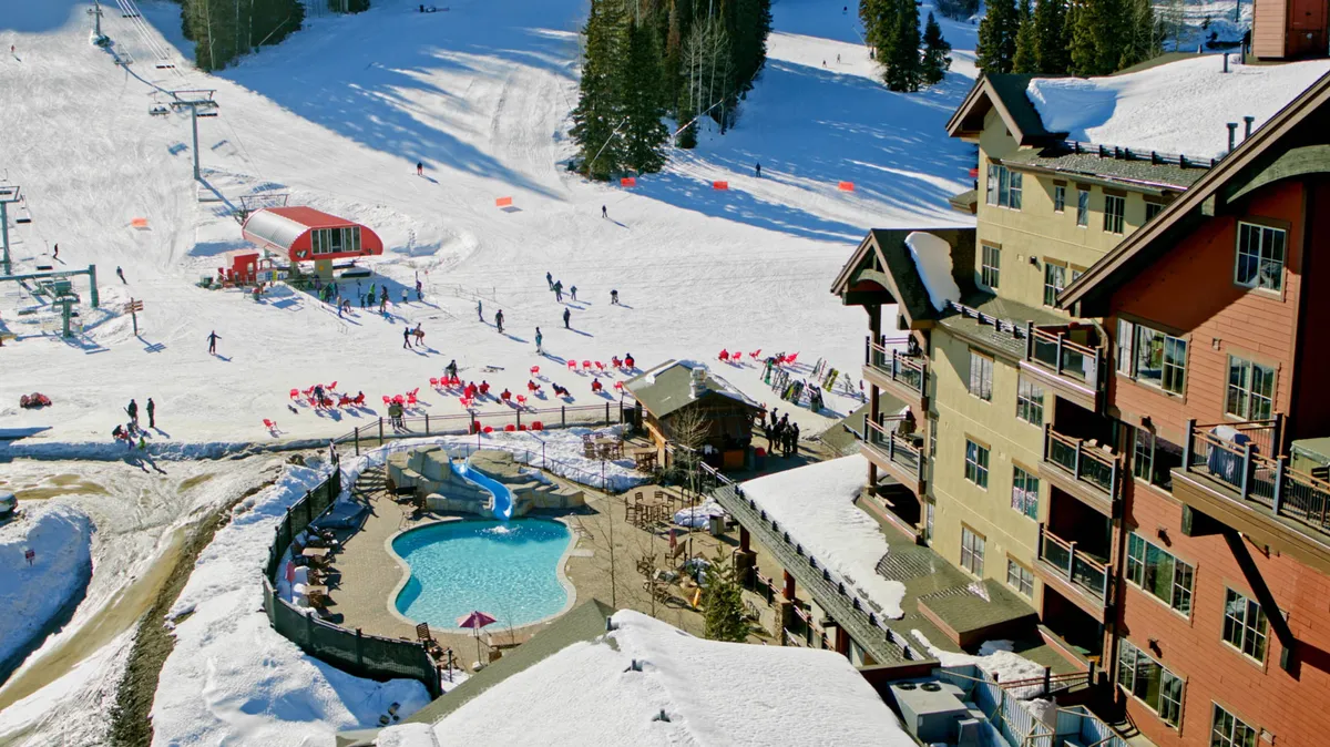 An exterior view showing ski slopes, a swimming pool, a ski lift, and nearby residential buildings, all surrounded by snow. The mood is energetic, suggesting active days outdoors followed by relaxation and socializing.