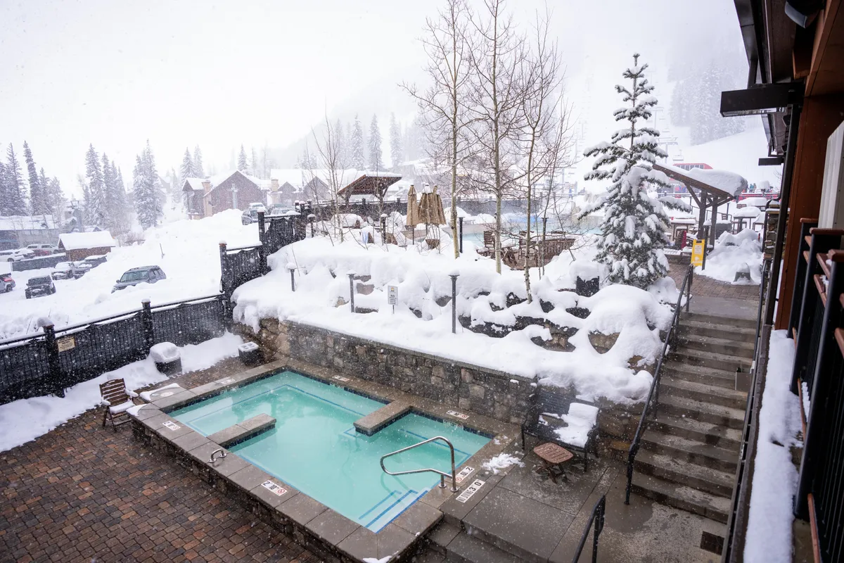 A tranquil winter scene featuring a heated outdoor pool or hot tub set amid snow-covered landscapes and tall evergreens. The inviting water contrasts beautifully with the snowy surroundings, evoking relaxation and comfort.