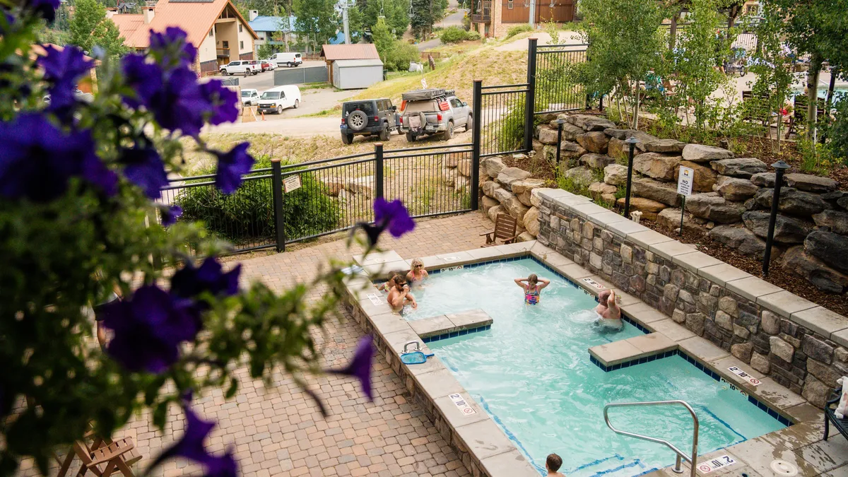 A bright, lively scene showing a swimming pool bordered by flowering plants and greenery, with people relaxing and socializing in and around the water. The mood is leisurely and social, ideal for family fun.