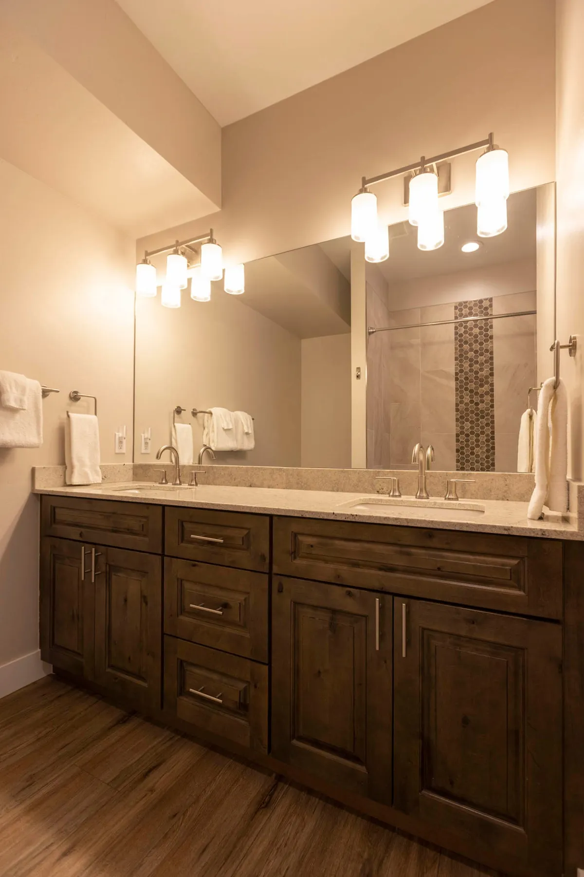 A modern bathroom featuring a double sink vanity, elegant light fixtures, a large mirror, and a spacious shower area. The serene setting is designed for personal grooming and relaxation, offering a spa-like experience.