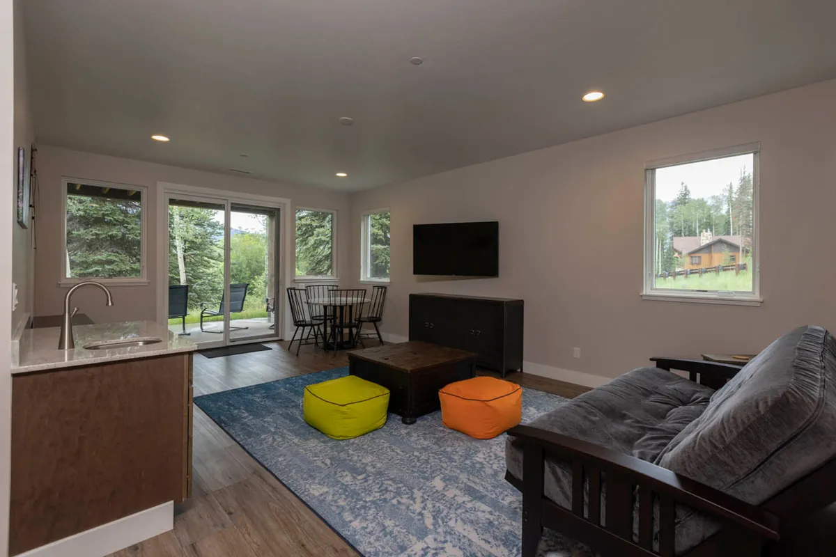 A spacious, sunlit living and dining area with a comfortable sofa, colorful ottomans, a large dining table, and expansive windows that invite natural light and display beautiful outdoor views. The space is arranged for both relaxation and communal activities like game nights or meals.