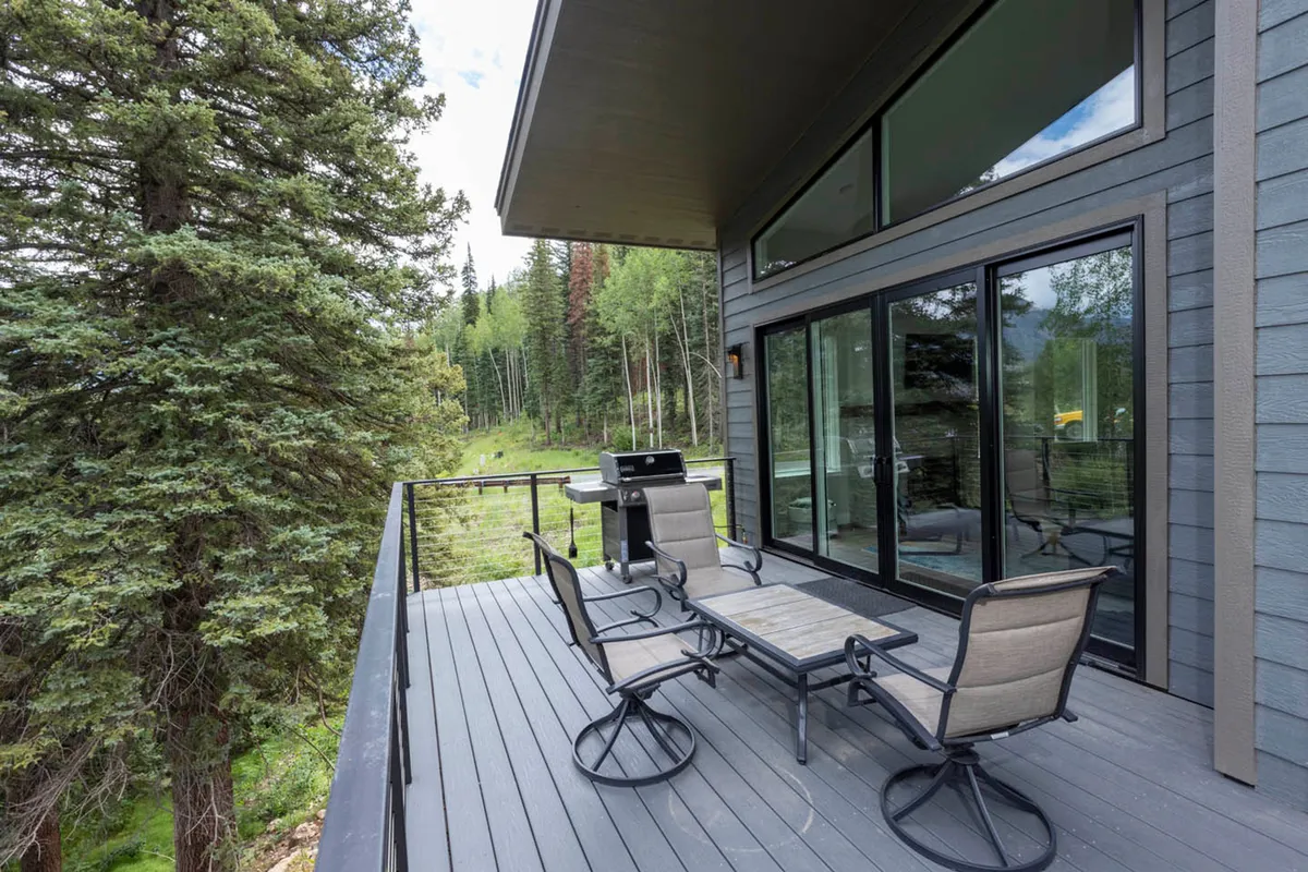 A peaceful exterior seating area with a grill and comfortable chairs sits just outside large glass doors, nestled amongst trees. The space offers tranquil outdoor dining or relaxing moments surrounded by nature, with hints of scenic views beyond.