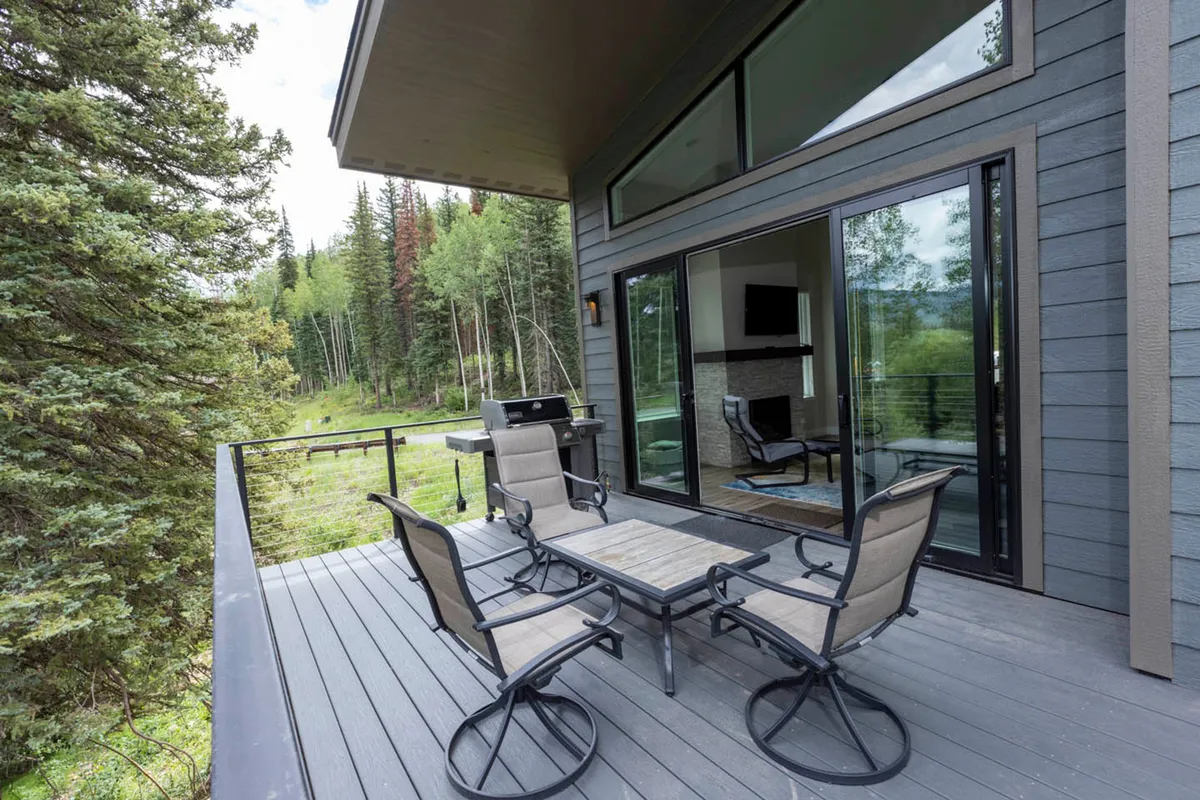 A spacious deck furnished with comfortable seating, a grill, and surrounded by lush trees. Large glass windows hint at the inviting interior, while the deck is perfectly positioned for outdoor dining or simply enjoying nature views.