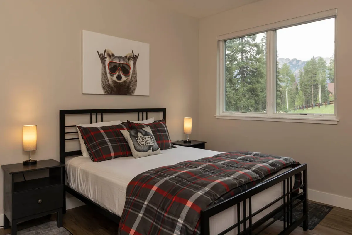A cheerful bedroom with a bed dressed in plaid bedding, a framed raccoon artwork above the headboard, nightstands with lamps, and a window offering a scenic view. The space is playful, inviting, and perfect for restful nights.